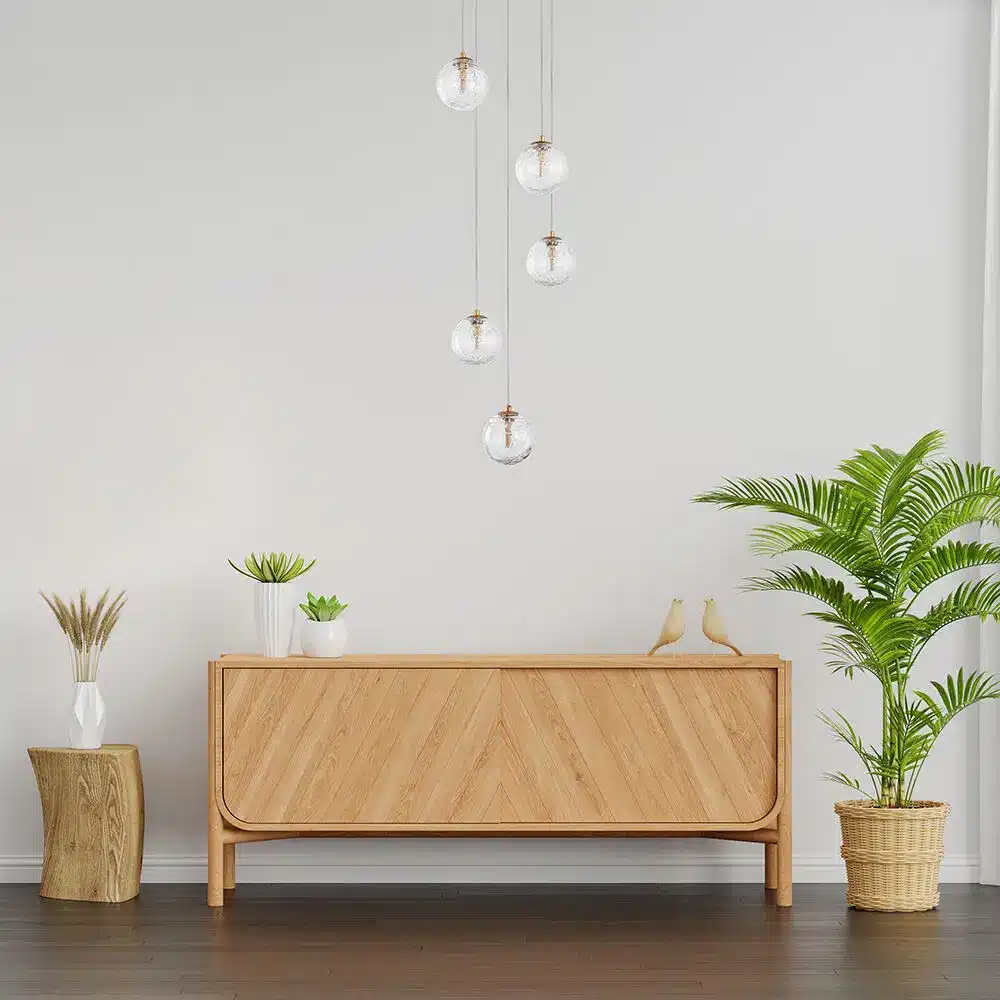 Suspension lumineuse moderne à pampilles en verre au-dessus d’un buffet en bois clair, plante verte et décor minimaliste. Suspension lumineuse moderne à pampilles en verre au-dessus d’un buffet en bois clair, plante verte et décor minimaliste.