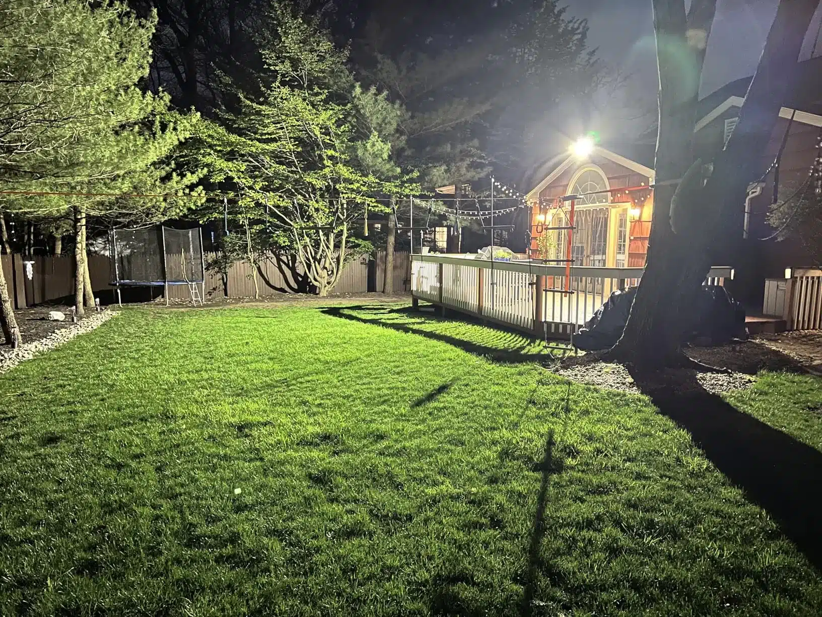 Gazon vert éclairé nocturne autour d’une maison avec terrasse, arbres, balançoire et trampo, éclairage extérieur puissant illuminant le jardin.