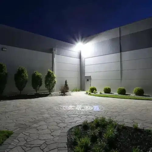 Allée circulaire pavée devant un mur de bâtiment, éclairée par une lampe extérieure au sommet, avec arbustes et parterres décoratifs.