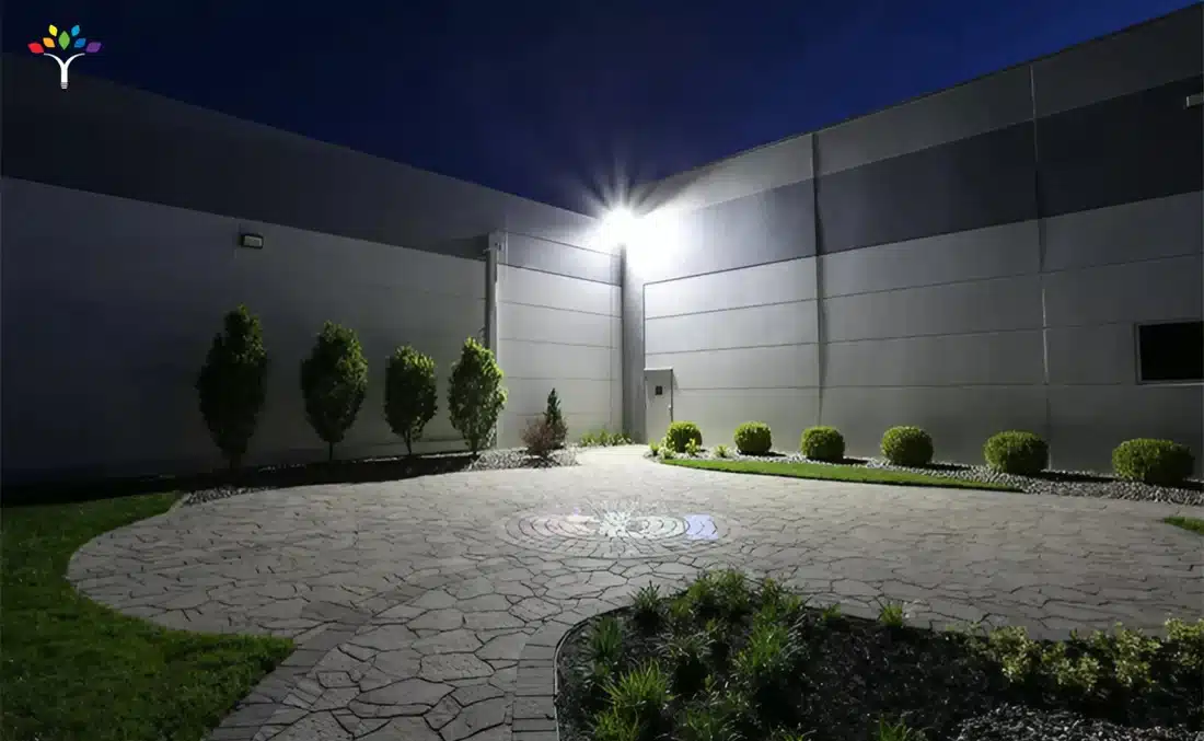 Allée circulaire pavée devant un mur de bâtiment, éclairée par une lampe extérieure au sommet, avec arbustes et parterres décoratifs.
