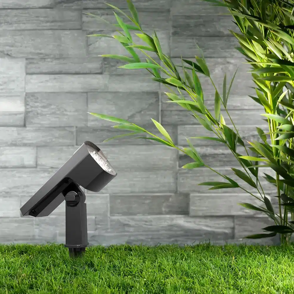 Projecteur extérieur LED noir orientable sur socle, installé sur gazon devant mur en pierre.