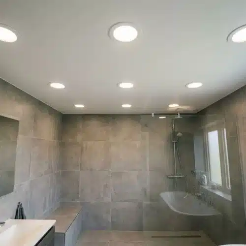 Salle de bains moderne éclairée par plusieurs spots encastrés, murs gris et douche à l’italienne en verre, fenêtre et espaces pratiques. Intègre le thème « comment positionner des spots en salle de bain ».