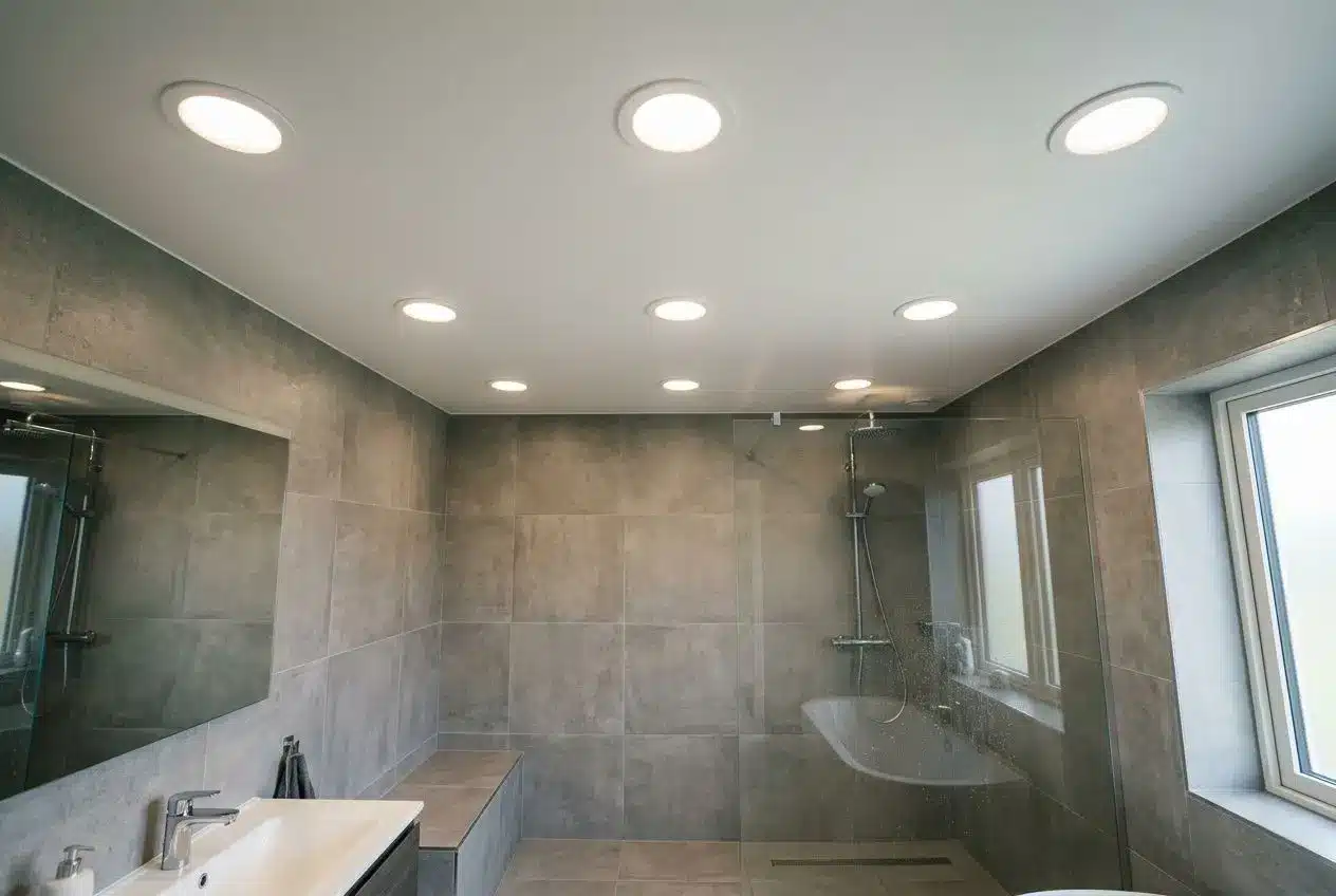 Salle de bains moderne éclairée par plusieurs spots encastrés, murs gris et douche à l’italienne en verre, fenêtre et espaces pratiques. Intègre le thème « comment positionner des spots en salle de bain ».