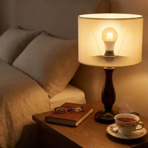 Lampe de chevet avec ampoule LED blanc chaud éclairant une table de nuit près du lit, tasse de café et livre posé.